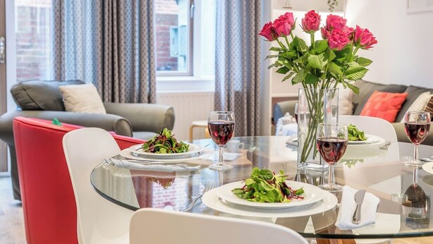 Un coin repas avec une table en verre dressée pour deux personnes, un vase rempli de roses roses et une vue sur le paysage urbain, visible à travers de grandes fenêtres.
