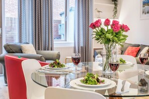 A dining area with a glass table set for two, a vase of pink roses, and a view of a cityscape through large windows. - East Mews Cottage, Southwold (Southwold)