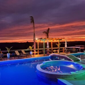 A pool area with lounge chairs and a hot tub, surrounded by a wooden pergola with string lights, under a sunset sky. - Chácara Arena JK (Ibiúna)