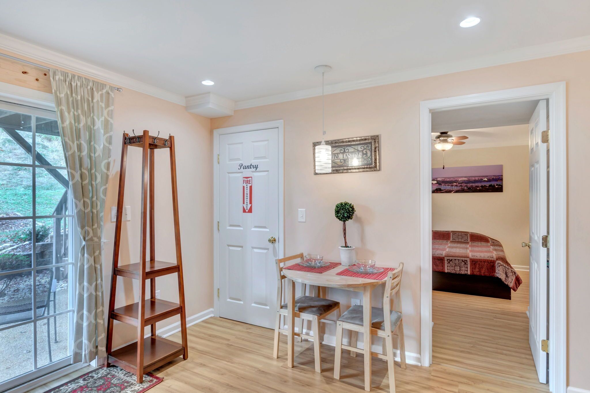A room with a dining table, chairs, a small potted plant, a bed with a patterned bedspread, and a door labeled "Pantry."
