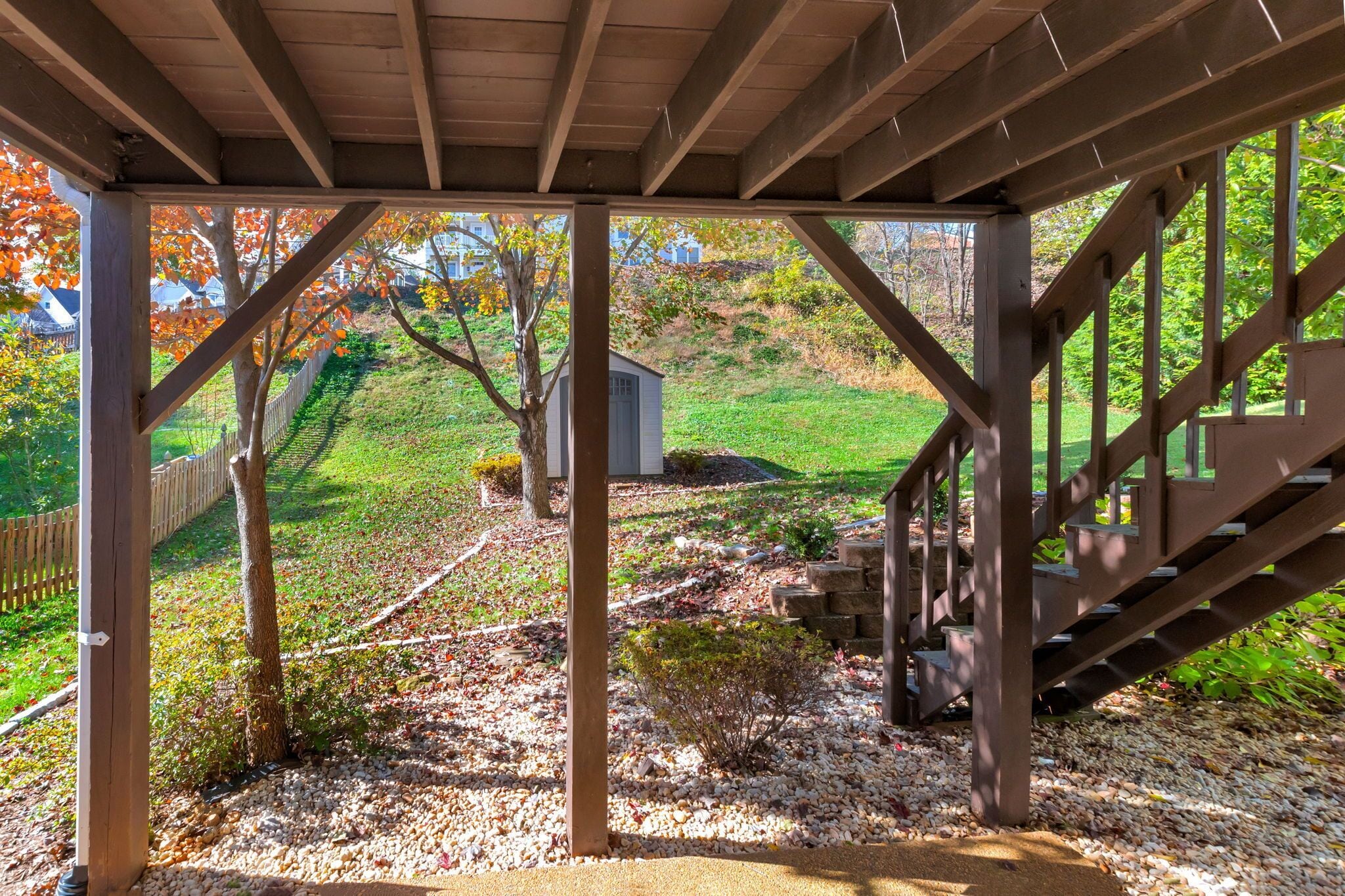 A covered outdoor area with a wooden staircase, a gravel path, and a view of a house and trees.