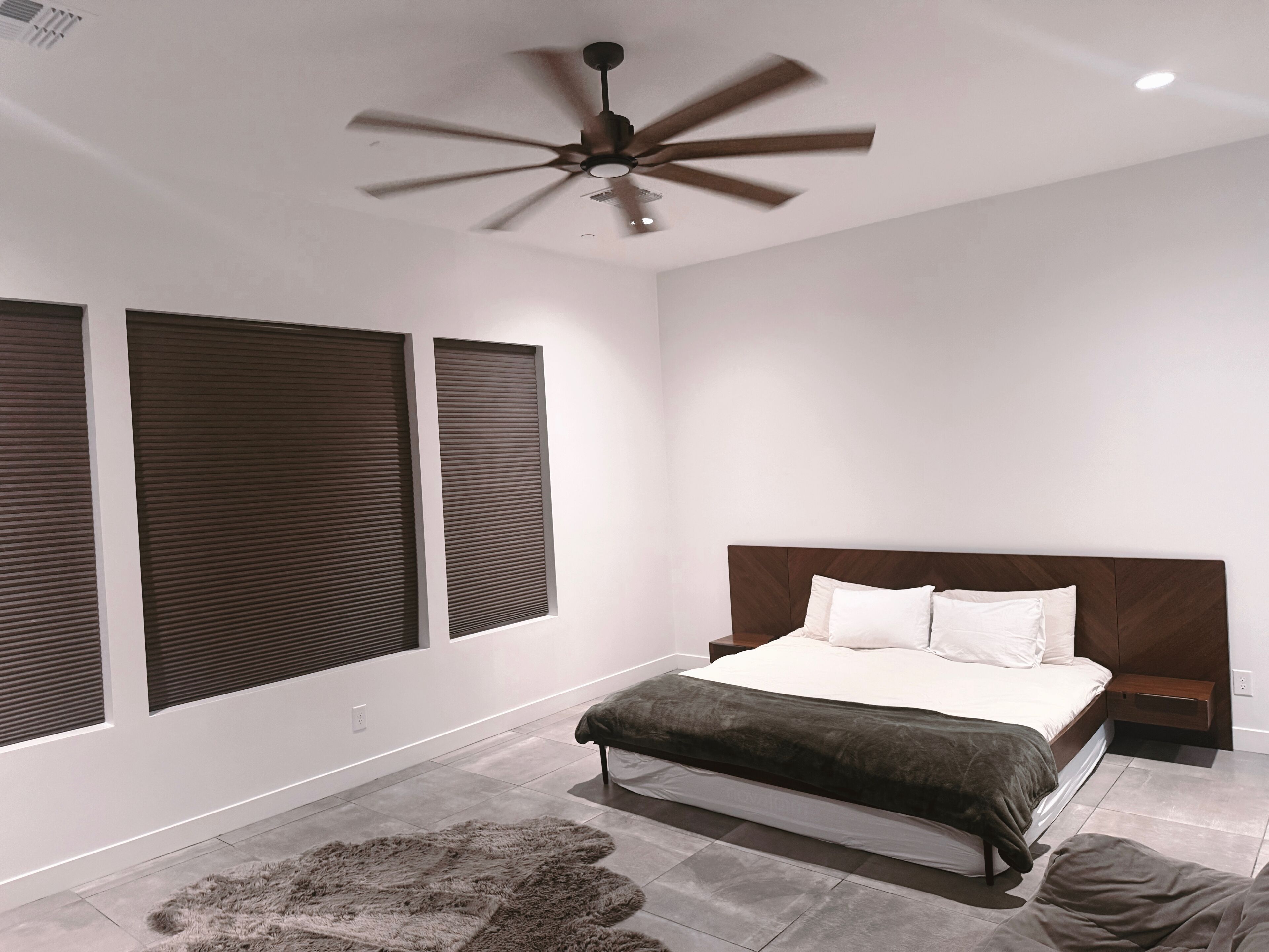 A modern bedroom with a large bed, a ceiling fan, and two windows with blinds.