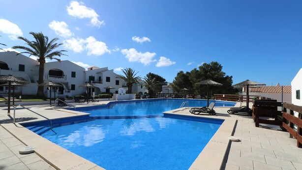 Piscina com espreguiçadeiras e guarda-sóis, cercada por edifícios brancos e palmeiras.