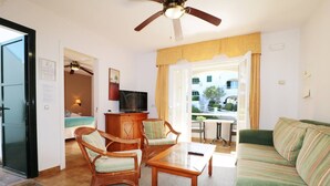 Uma sala de estar bem iluminada com ventilador de teto, um sofá verde, duas cadeiras de madeira, uma mesa de centro de madeira e uma TV em um móvel.