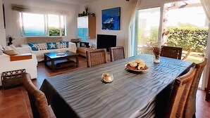 Ein Essbereich mit einem Tisch für zwei Personen, umgeben von Holzstühlen. Im Hintergrund befindet sich eine Couch und ein Fernseher.