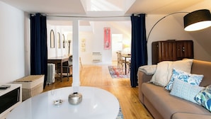 Un salón moderno con una mesa de centro blanca, un sofá color beige con cojines estampados, una lámpara de pie oscura y vistas al área de comedor a través de una puerta de cristal.
