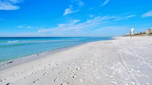 Une plage avec des empreintes de pas dans le sable, une eau d’un bleu limpide et des bâtiments en arrière-plan.