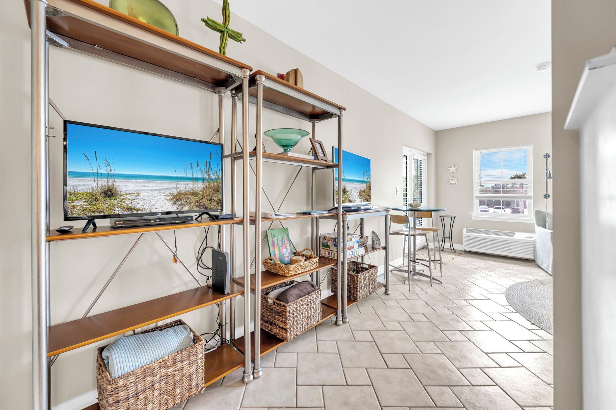Ein modernes Wohnzimmer mit einem großen Flachbildfernseher, einem Metallregal, Weidenkörben und Blick auf den Strand durch das Fenster.