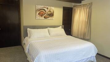 Een netjes opgemaakt bed met een witte spreidek en kussens, een donker houten deur, een abstract kunstwerk aan de muur en een raam met beige gordijnen.