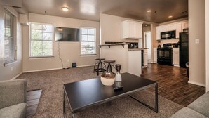 A spacious living area with a dark coffee table, a TV mounted on the wall, and a kitchen area with white cabinets and modern appliances. - Modern 1 Bed - Gated - Reserved Parking (Oklahoma City)