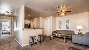 A spacious living area with a sofa, coffee table, and two bar stools. A ceiling fan and artwork are visible on the walls. - Modern 1 Bed - Gated - Reserved Parking (Oklahoma City)