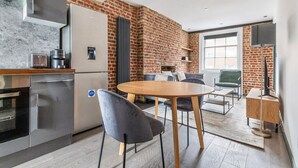 Eine moderne Küche mit Backsteinwand, ein Essbereich mit rundem Tisch und Stühlen sowie ein Wohnbereich mit Sofa und Fernseher.