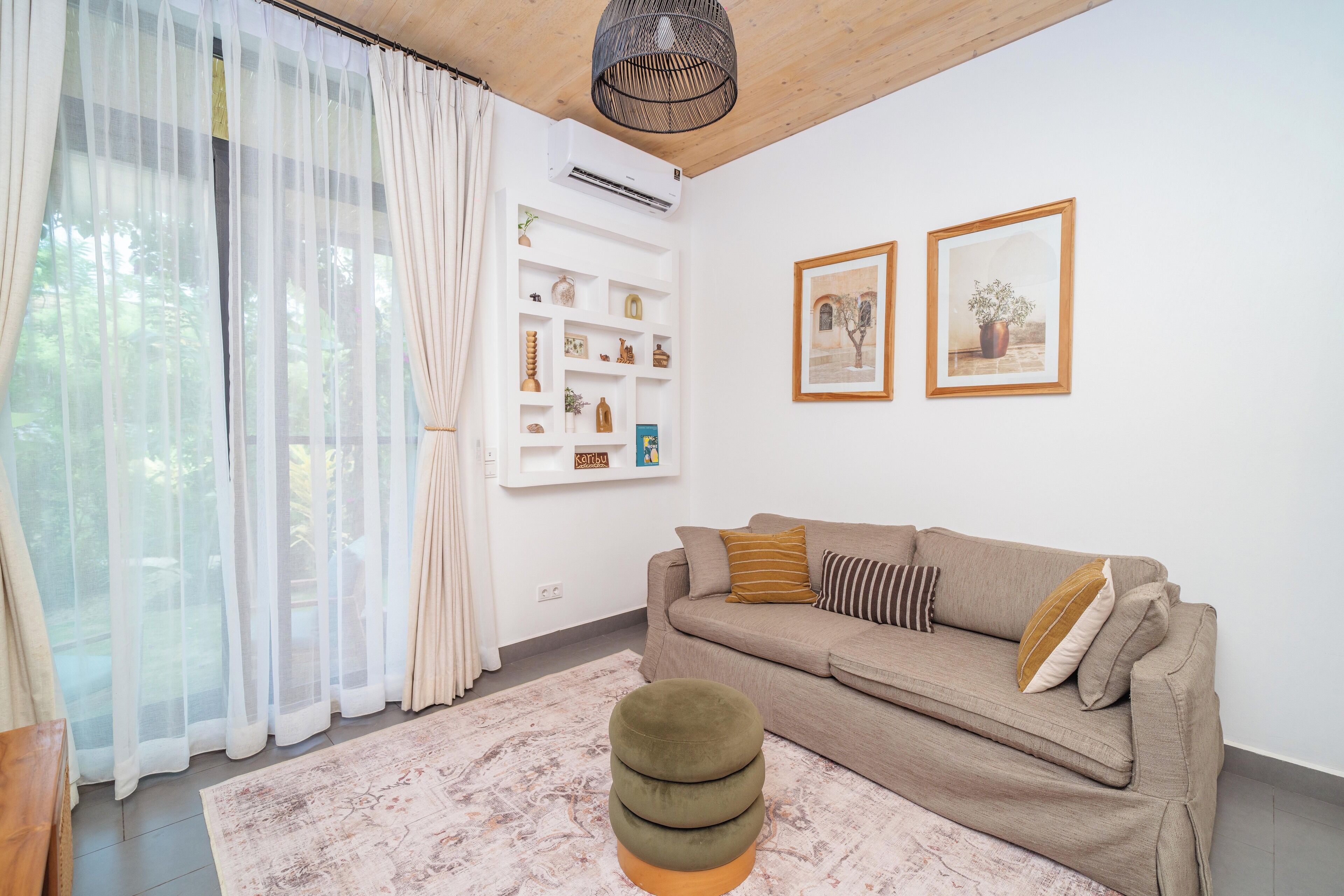 Un soggiorno con un divano color beige, un poggiapiedi rotondo, un condizionatore d'aria bianco e due quadri appesi al muro.