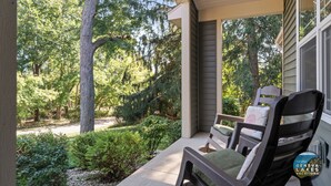 A covered porch with a wooden chair and a view of a wooded area.