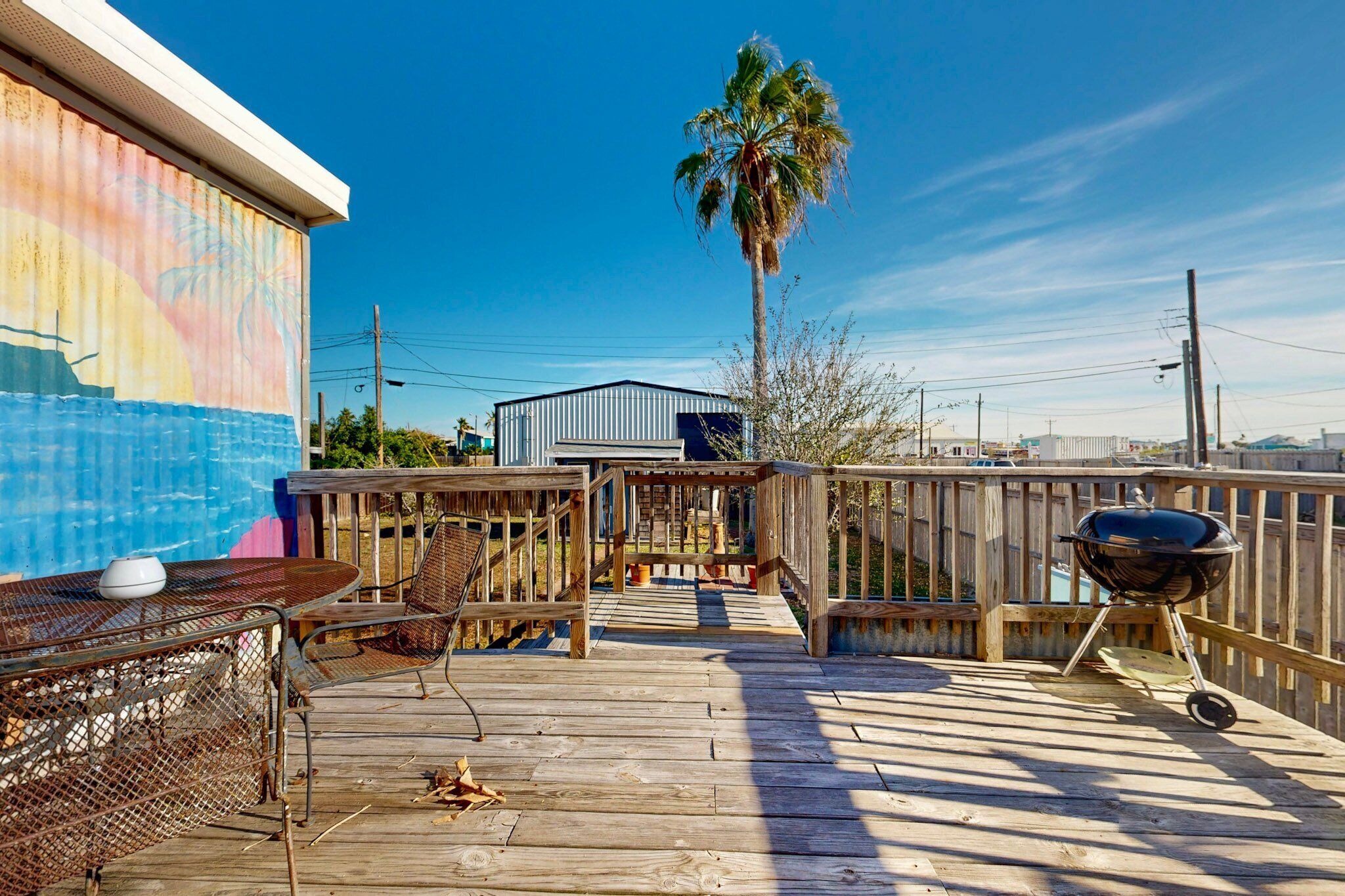 Una terraza de madera con sillas de metal y una parrilla, con vista a un cielo azul y una palmera.