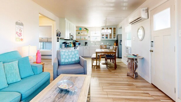 A spacious living room with a wooden coffee table, a blue sofa, and a ceiling fan. A kitchen with white cabinets and a dining area with a wooden table and chairs is visible in the background. - Pet Friendly, Golf Cart Accessible, Old Town Port A (Port Aransas)