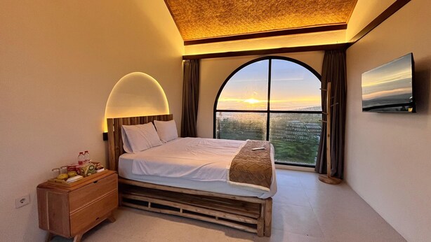 Een slaapkamer met een groot bed, nachtkastje, televisie en uitzicht op de ondergaande zon door een groot raam.