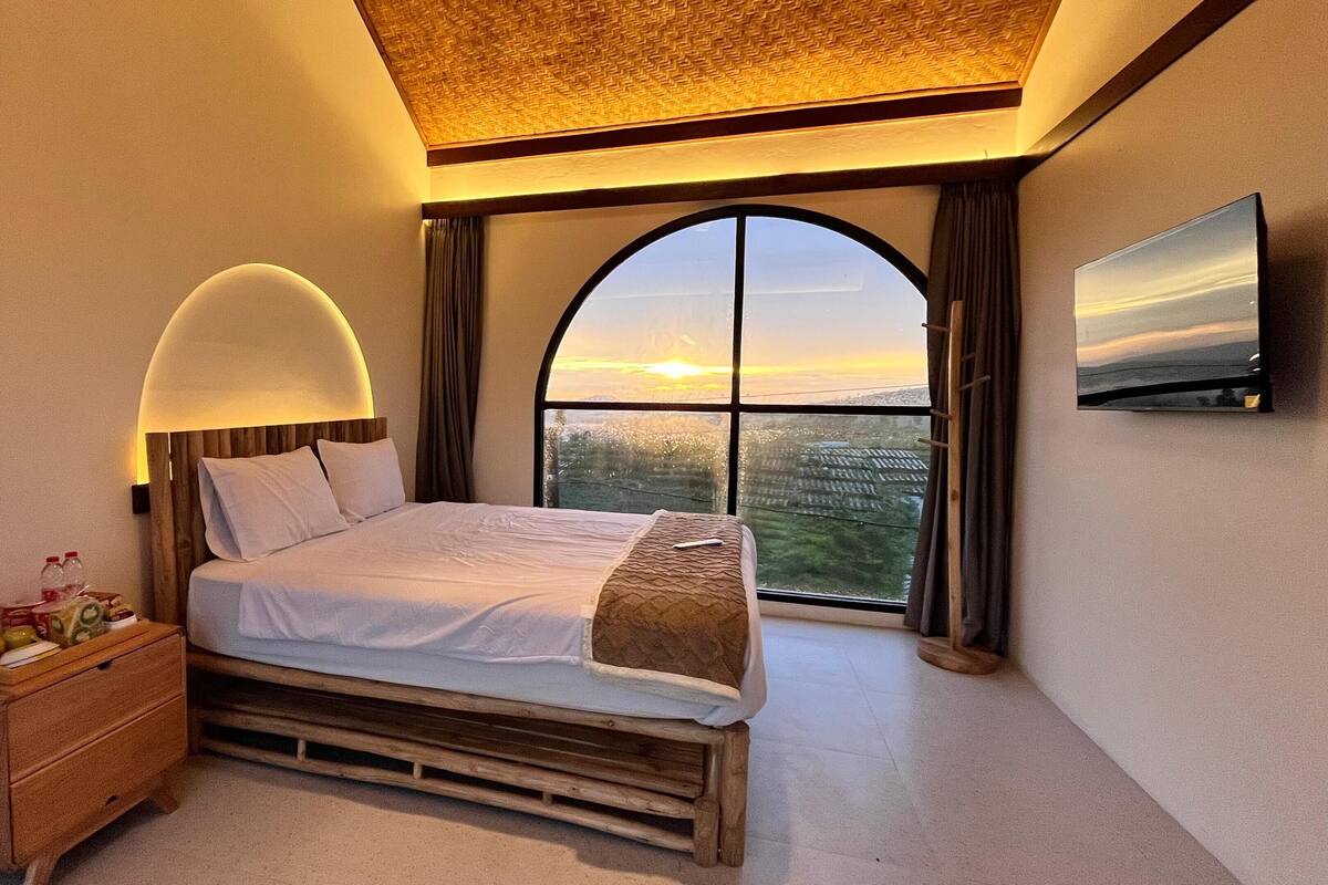 A bedroom with a large bed, a nightstand, a TV, and a view of the sunset through a large window.