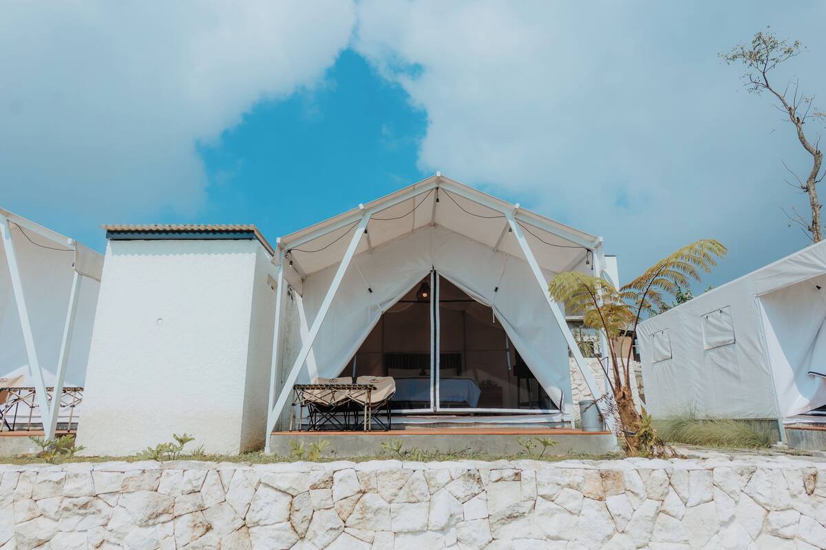 A modern tent-style accommodation with a stone wall in front, a bed inside, and a small table and chair outside.