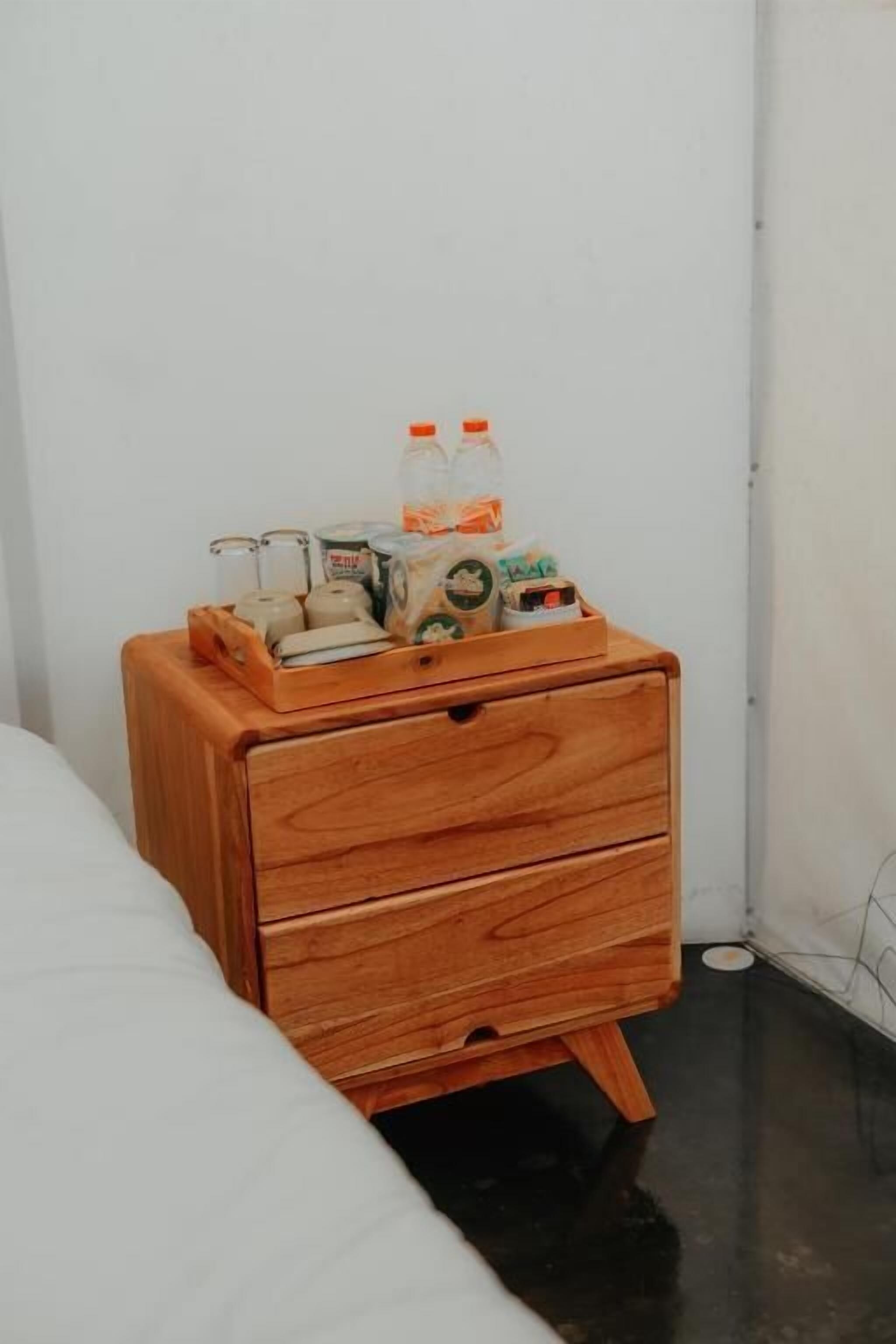 A wooden bedside table with a drawer, a tray holding various items including bottles and containers, against a white wall in a room with a black floor.