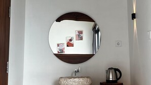 Een houten wastafelmeubel met een ronde spiegel, een wastafel en een waterkoker op een aanrecht.