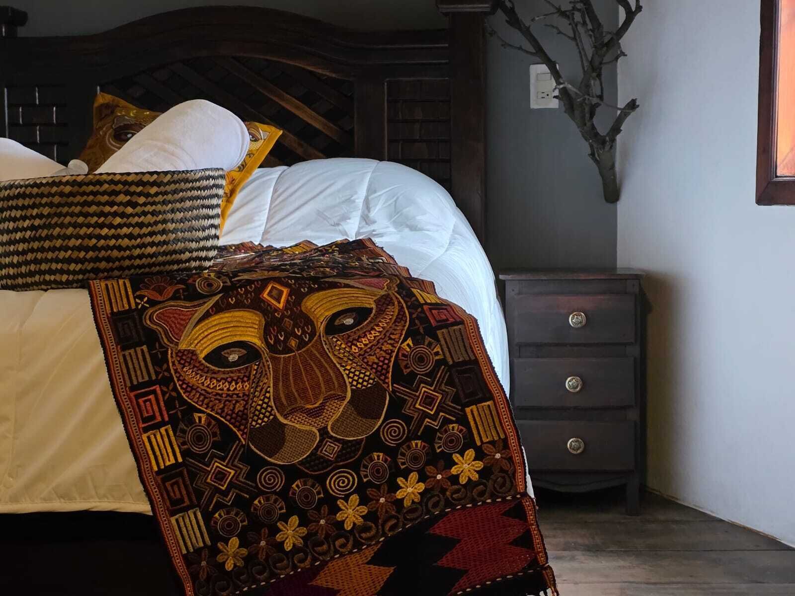 Una cama bien hecha con sábanas blancas y una manta estampada. Una mesita de noche de madera con un florero de ramas está al lado de la cama.