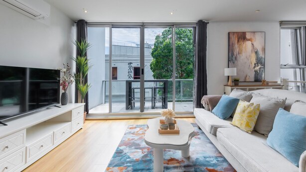 A modern living room with a large TV, a sofa with cushions, a coffee table, and a view of the outdoors through large windows. - Loft Life Footscray Near Cafes & CBD (Footscray)