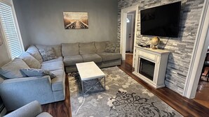 A modern living room with a sectional sofa, a flat-screen TV mounted on a stone wall, a fireplace, and a patterned rug. - Charming 4-bedroom house in lovely Scranton perfect for families (Scranton)