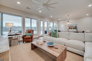 Ein geräumiges Wohnzimmer mit großem Fenster, Deckenventilator, Couchtisch aus Holz, einer Couch und Blick aufs Wasser.