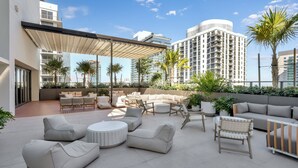 A rooftop patio with outdoor seating, palm trees, and modern buildings in the background. - The Crosby Luxury Corner 2BR/2BA Rooftop Pool & GYM | 1614 (Miami)