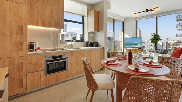 A modern kitchen with wooden cabinets, a dining area with wicker chairs, and a view of the city and ocean. - The Crosby Luxury Corner 2BR/2BA Rooftop Pool & GYM | 1614 (Miami)