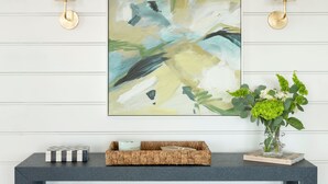 Een moderne woonkamer met een donkere console tafel, twee gestreepte poefs, een groot abstract schilderij en een vaas met groene planten.