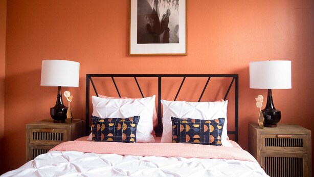 Una cama bien hecha con ropa de cama blanca y rosa, dos mesitas de noche con lámparas y un cuadro enmarcado en la pared.