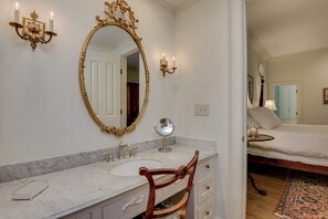 Un baño con lavamanos de mármol, un espejo grande y decorativo, y una silla de madera.