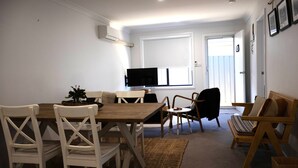 Een eethoek met een houten tafel en witte stoelen, een televisie aan de muur en een raam met blinds.