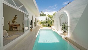 A modern pool area with a clear blue pool, white walls, and a wooden table with chairs. - Villa Marysol Uluwatu (Uluwatu)