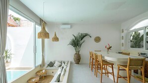 A modern living room with a large window, a sofa, a dining area with wooden chairs, and a potted plant. - Villa Marysol Uluwatu (Uluwatu)