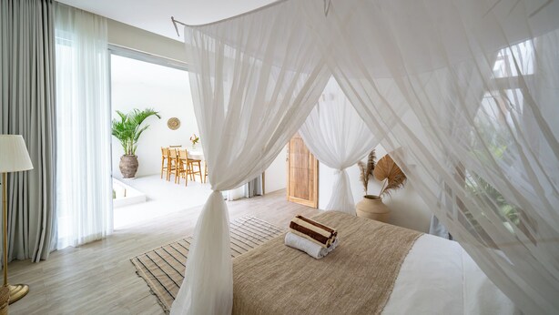 A spacious bedroom with a large bed, sheer curtains, and a view of a dining area. - Villa Marysol Uluwatu (Uluwatu)