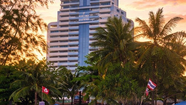 Un hotel de gran altura con varios pisos, ubicación frente a la playa y la bandera de Tailandia.