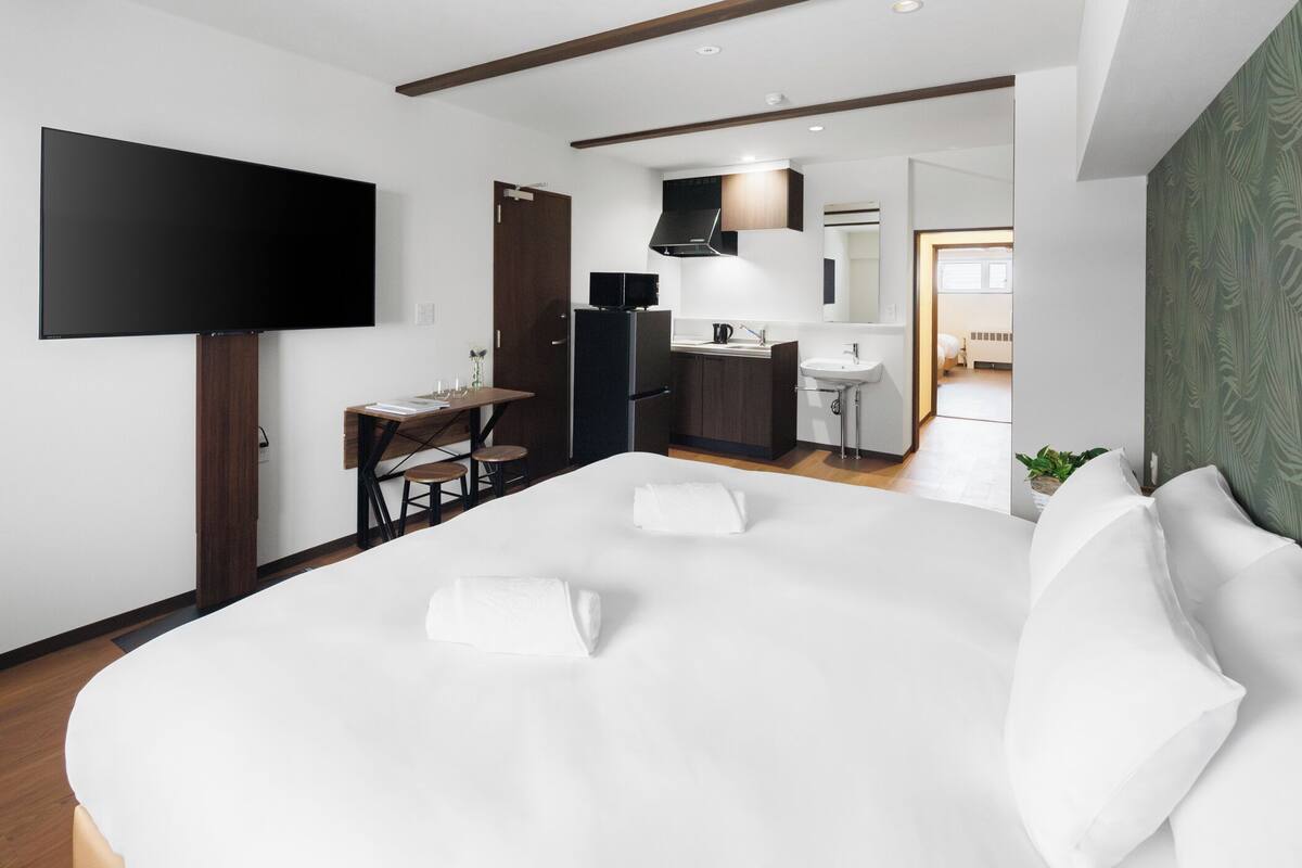 A modern hotel room with a large bed, a flat-screen TV, a small kitchen area, and a bathroom visible through an open door.