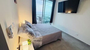 Ein Schlafzimmer mit einem Bett, einem Nachttisch mit Lampe, einem an der Wand befestigten Fernseher und Meerblick aus dem Fenster.