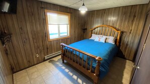 A bedroom with wooden walls, a bed with a blue bedspread, a wooden headboard, a window with blinds, and a ceiling light. - Peaceful Country Getaway Near Dillon, MT (Dillon)