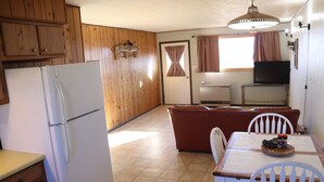 A compact kitchen with wooden cabinets, a white refrigerator, a dining table with chairs, and a television in the background. - Peaceful Country Getaway Near Dillon, MT (Dillon)