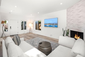 Un salon moderne avec un canapé blanc, une table basse ronde et une cheminée. Il y a deux fenêtres avec des rideaux, un lampadaire et un tableau représentant un paysage côtier.