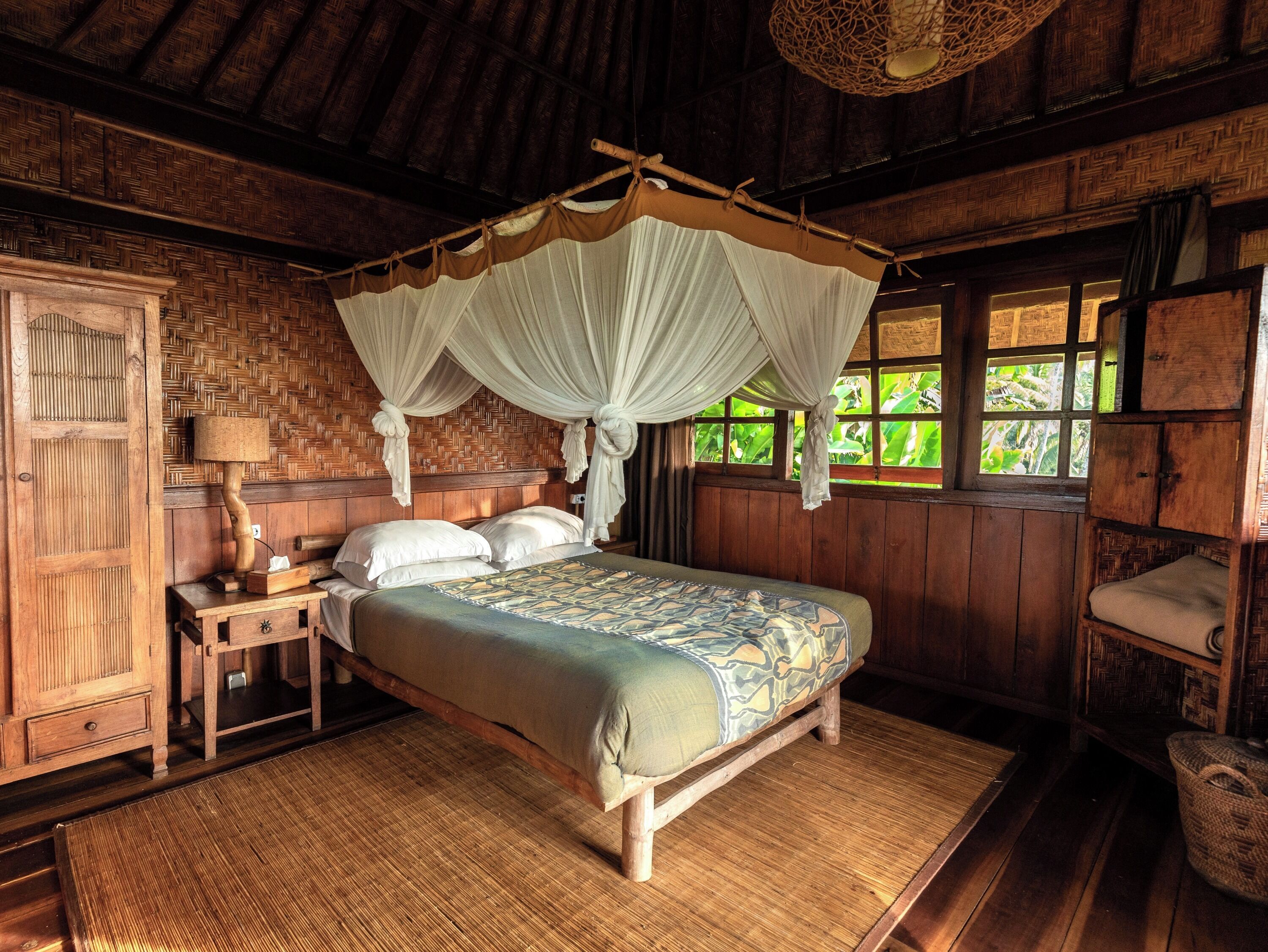A traditional wooden room with a four-poster bed, a woven mat on the floor, and a window with a view of greenery.