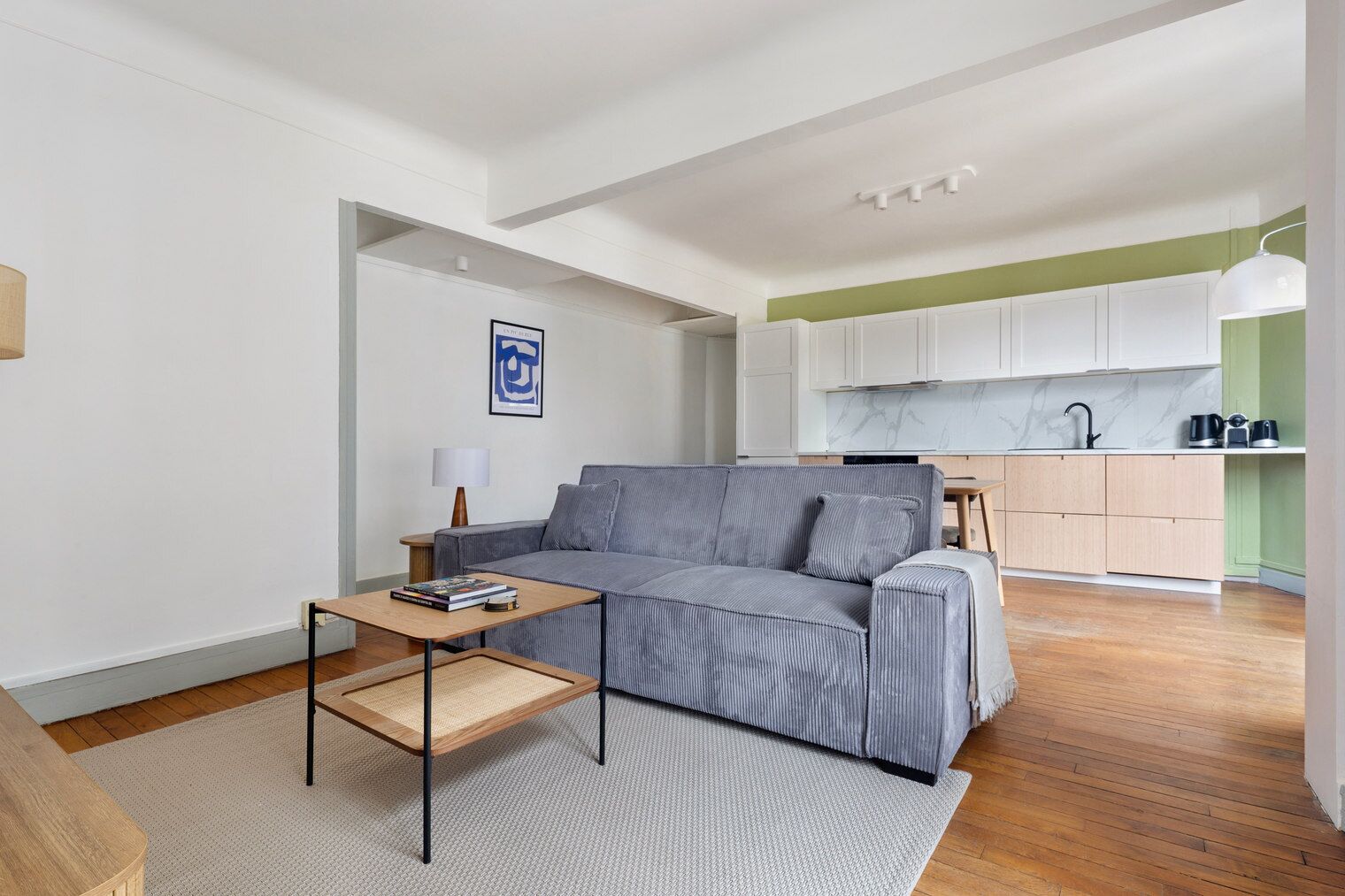 A modern living room with a grey sofa, a small wooden coffee table, and a white rug. A kitchen with white cabinets and a green accent wall is visible in the background.