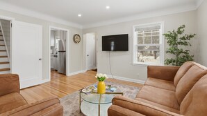 A living room with a brown sofa, a glass coffee table with a yellow vase, a flat-screen TV mounted on the wall, and a window with a view of the outdoors. - Charming 4BR Home; 3 Levels; Near Airport & MOA (Saint Paul)