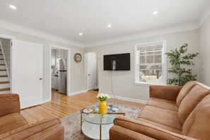 A living room with a brown sofa, a glass coffee table with a yellow vase, a flat-screen TV mounted on the wall, and a window with a view of the outdoors. - Charming 4BR Home; 3 Levels; Near Airport & MOA (Saint Paul)