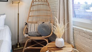 Ein Zimmer mit einem Korbstuhl, einem kleinen runden Tisch mit einer Vase, einer Stehlampe und einem Bett.