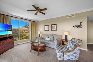 A hotel room with a large window, a ceiling fan, a sofa, a coffee table, a TV, and decorative wall art. - Honua Kai | Ocean View 5 BR, Sleeps 16 | Car Incl. w/ 6+ Nights | HKK ML-3559 by KBM (Lahaina)